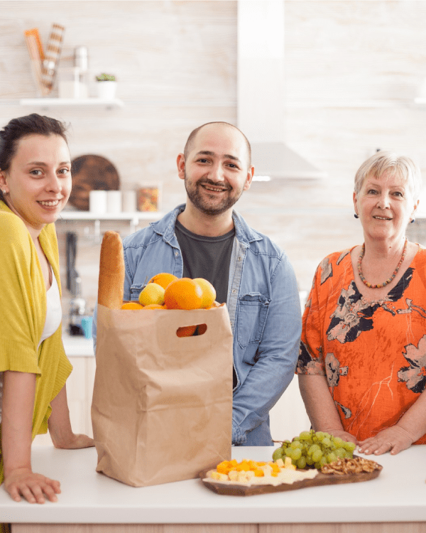Four people around a kitchen counter with a grocery bag and fresh fruit.