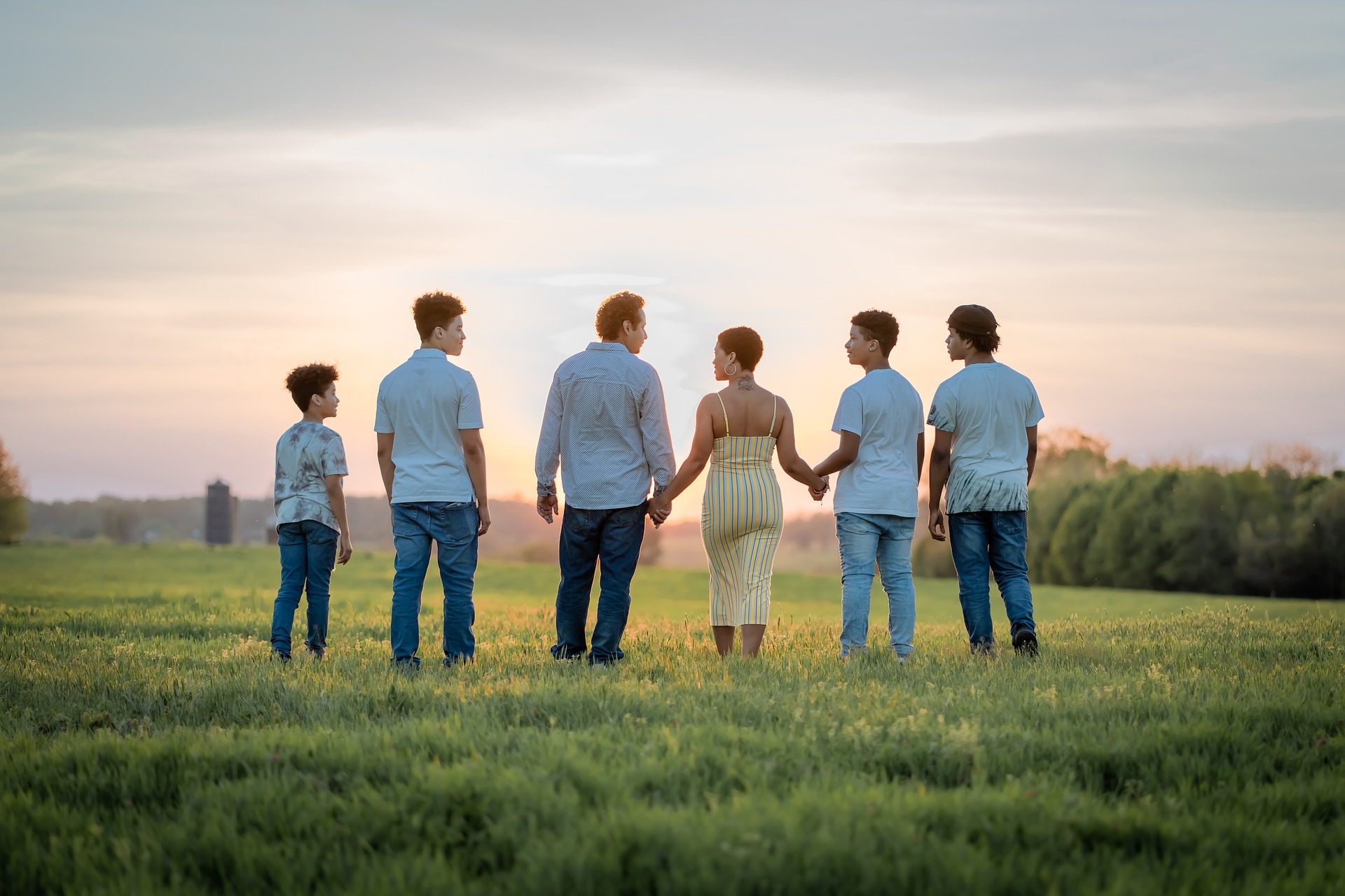 Group of people holding hands, facing sunset in a field.